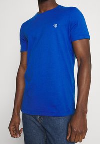 Blaues T-Shirt aus Baumwolle mit Rundhalsausschnitt und kurzen Ärmeln. Ein kleines weißes Logo ist auf der linken Brust aufgedruckt.