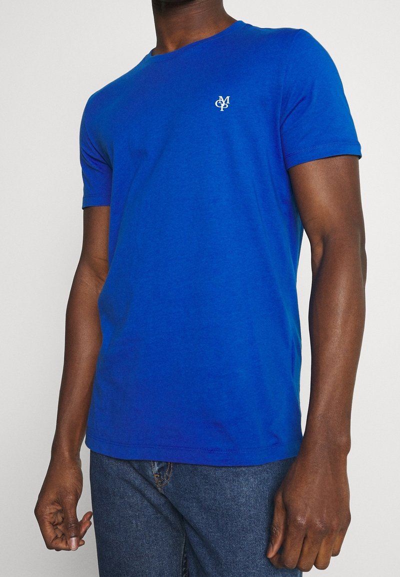 Blaues T-Shirt aus Baumwolle mit Rundhalsausschnitt und kurzen Ärmeln. Ein kleines weißes Logo ist auf der linken Brust aufgedruckt.