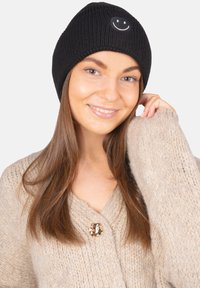 Bonnet noir en maille côtelée avec un petit visage souriant blanc. Le modèle porte un cardigan en maille beige oversized.