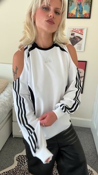 Jauns cilvēks ar blondiem matiem, tērpies baltā Adidas auksto plecu topā un melnos šortos, stāv telpā ar ietērptām sienas gleznām.