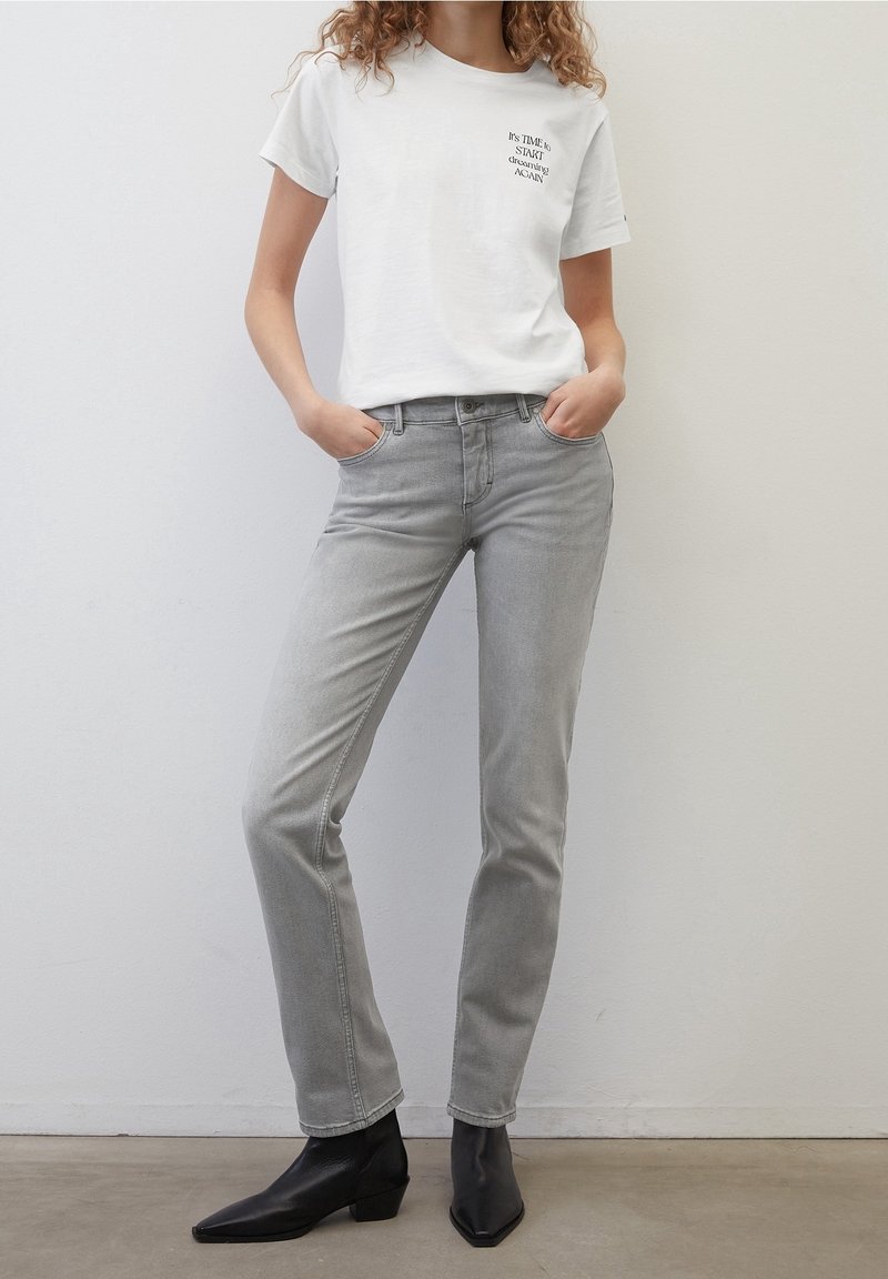 Camiseta blanca con estampado de texto, combinada con unos pantalones vaqueros gris claro y botines negros. Diseño ajustado con detalles sutiles y texturas suaves.