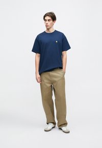 Modra bombažna majica s kratkimi rokavi in krožnim izrezom ter majhnim zlatim logotipom. Pairs z bež širokimi hlačami in belimi supergami.