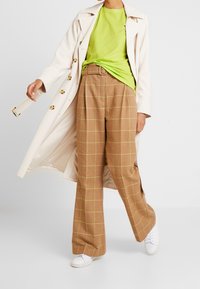 Beige rutede vide bukser med gule linjer, parret med en lys grøn t-shirt og en lang hvid frakke. Hvide sneakers giver kontrast.