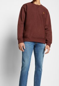 Brun sweatshirt med ribbade ärmslut och nederkant, med en liten logotyp på bröstet. Bärs över ljusblå jeans. Mjuk struktur, avslappnad passform.