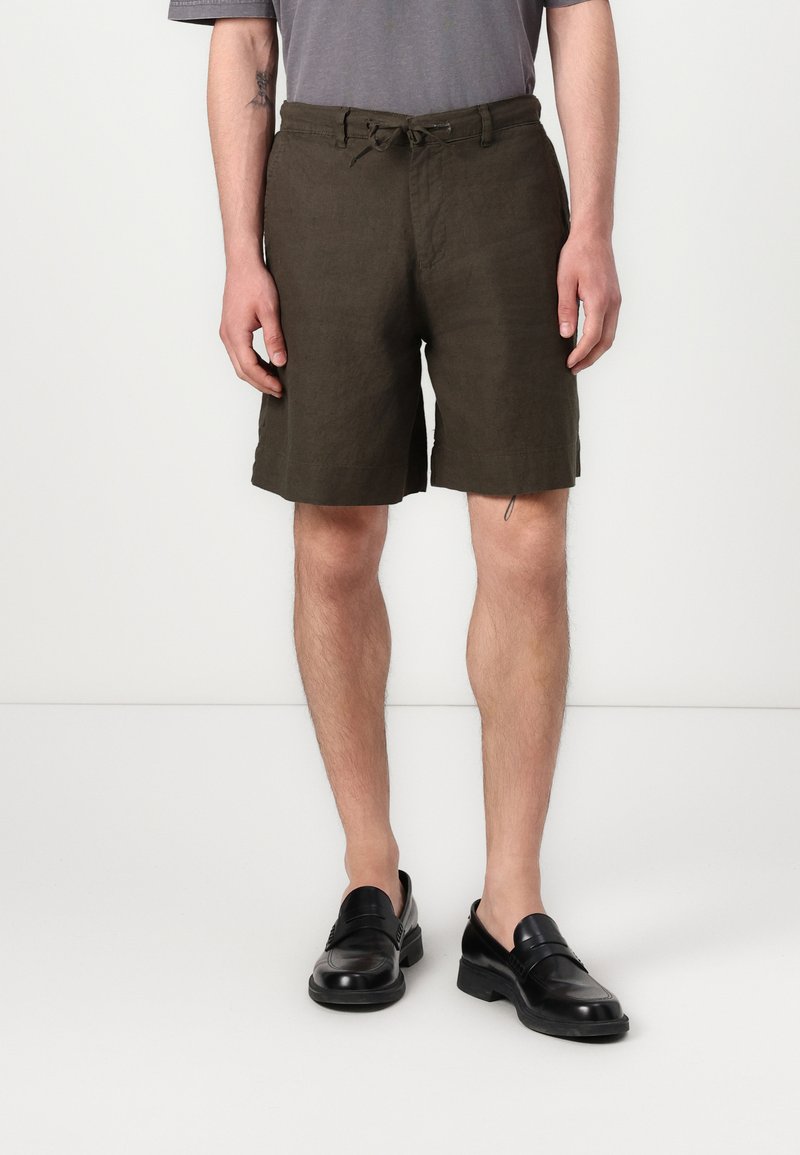Shorts en lin vert olive avec une taille élastique et un cordon de serrage, présentant des fentes latérales et une coupe décontractée, associés à des chaussures noires.