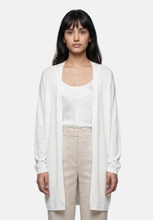 Vrouw met lang zwart haar die een witte open cardigan, een wit topje en beige hoge taille broek draagt, staand tegen een effen achtergrond.