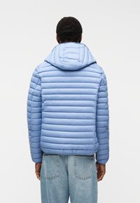 Άτομο που φοράει ανοιχτό μπλε καπιτονέ μπουφάν με κουκούλα και φαρδιά μπλε τζιν, στεκόμενο με την πλάτη προς τα έξω σε απλό φόντο.