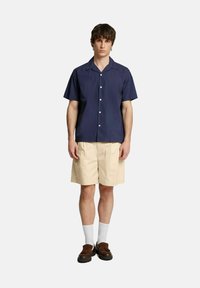 Camicia blu navy a maniche corte con vestibilità rilassata, abbinata a pantaloni beige. Scarpe marroni e calzini bianchi completano l'outfit, caratterizzato da un design casual.