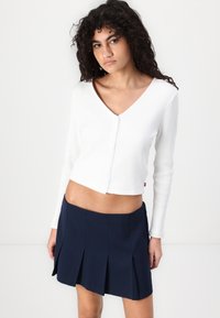 White ribbed long-sleeve cropped cardigan with a v-neck and button closure, paired with a navy pleated mini skirt.