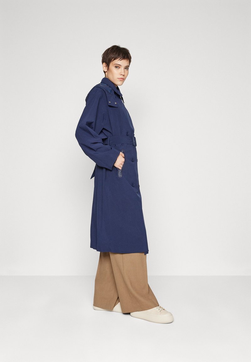 Personne portant un trench-coat bleu marine ceinturé, un pantalon large beige et des baskets blanches, se tenant de côté avec une main dans la poche du manteau.