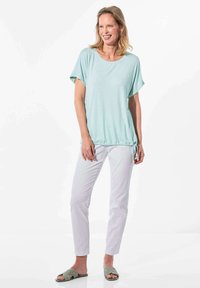 Lichtgroen kortemouwen top met een ronde halslijn en een koord onderaan, gecombineerd met witte slim-fit broek en grijze instapsandalen.