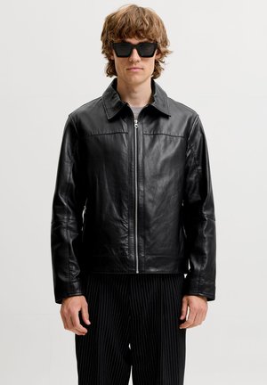 Junger Mann trägt schwarze Lederjacke mit Reißverschluss, schwarze Sonnenbrille und schwarze Nadelstreifenhose, steht vor schlichter hellem Hintergrund.