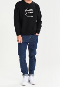 Černá mikina s bílým obrysem písmene "G", spárovaná s modrými džíny a bílými teniskami. Pohodlný střih a jednoduchý design.