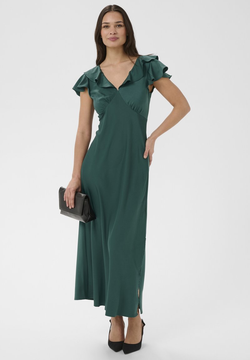 Femme en robe courte vert foncé à volants et manches courtes, tenant une pochette noire, portant des escarpins noirs à bout pointu, debout avec une main sur la hanche.