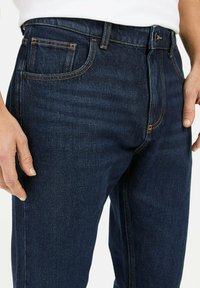 Dunkelblaue Jeans aus Denim mit einem klassischen Fünf-Taschen-Design, orangefarbenen Nähten und einer geraden Passform. Glatte Textur, Knopfverschluss.