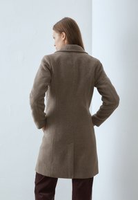 Manteau en mélange de laine marron avec un design structuré, col cranté et poches latérales. Caractérisé par des coutures verticales et une coupe légèrement cintrée.