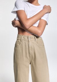 Pantalones de tiro alto en beige con cintura elástica y bolsillos frontales, combinados con una camiseta blanca de manga corta lisa. Textura de tela suave.