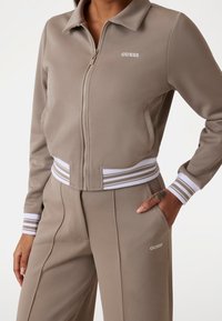 Beige trainingspak met bijpassend jack met ritssluiting en broek. Jack heeft witte gestreepte manchetten en het 'GUESS' logo; gestructureerde stof, losse pasvorm.