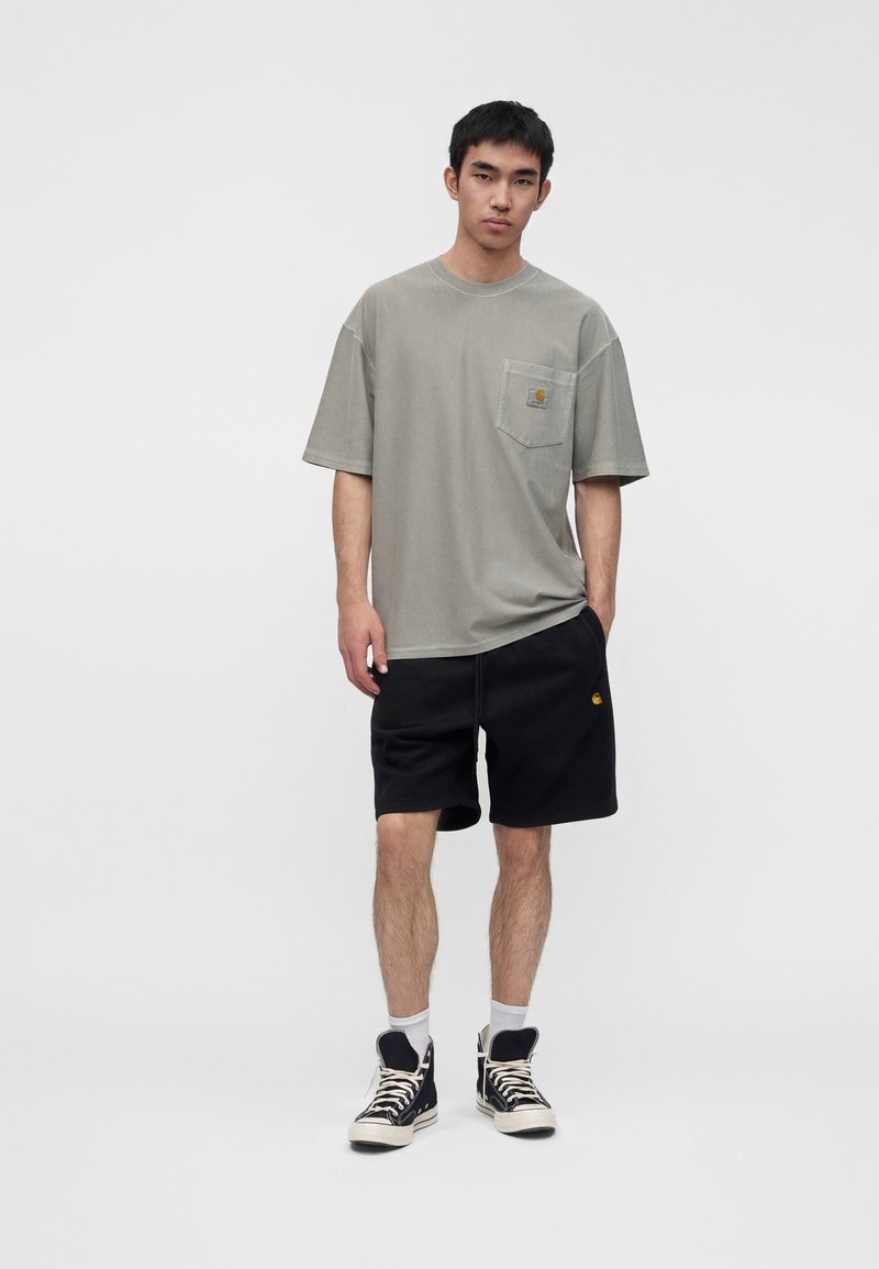 Ein junger Mann steht und trägt ein locker sitzendes graues T-Shirt mit Taschen, schwarze Shorts, weiße Socken und schwarze High-Top-Sneaker vor einem einfarbigen Hintergrund.