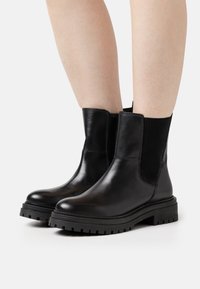 Botas de cuero negro hasta el tobillo con paneles laterales elásticos, suelas de goma gruesas y un acabado suave. Presenta una punta redondeada y una suela con relieve robusto.