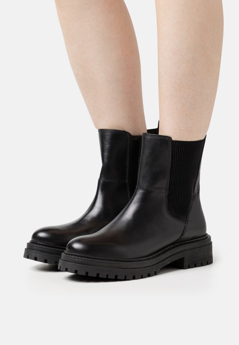 Botas de cuero negro hasta el tobillo con paneles laterales elásticos, suelas de goma gruesas y un acabado suave. Presenta una punta redondeada y una suela con relieve robusto.