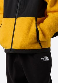 Φλις μπουφάν με κίτρινο σώμα και μαύρους ώμους, με τσέπες με φερμουάρ. Μαύρο παντελόνι φόρμας με λευκό λογότυπο στον μηρό.