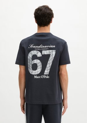 Άνδρας φοράει σκούρο μπλουζάκι με το κείμενο "Scandinavian Football Community 67 Marc O’Polo" και σχέδιο πλέγματος στην πλάτη, στέκεται μπροστά σε λευκό φόντο.