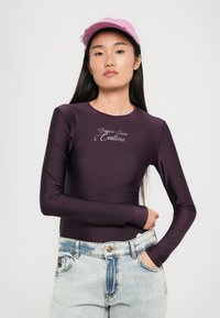 Fialový body s dlhým rukávom, okrúhlym výstrihom a strieborným logom "Versace Jeans Couture", doplnený svetloružovými džínovými šortkami.