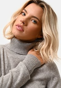 Szary sweter z golfem, z prążkowaną teksturą i mankietami. Zbliżenie na twarz modelki, z delikatnymi falami we włosach i neutralnym kolorem ust.