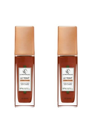 Deux bouteilles de fond de teint avec des bouchons carrés en cuivre métallique. Les récipients en verre sont mostly transparents, mettant en valeur un produit brun riche à l'intérieur. Les étiquettes affichent les détails de la marque et des ingrédients.