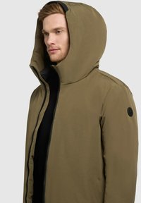 Olivgrön huva jacka med dragkedja, med en texturerad tyg och en svart logotyp patch på vänster ärm.