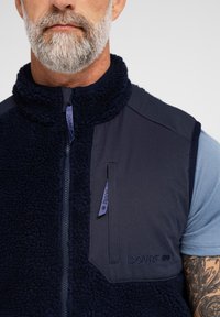 Marine fleece vest med høj krave, tekstureret krop og glatte skulderpaneler; har en lynlås lomme og diskret branding på højre side.