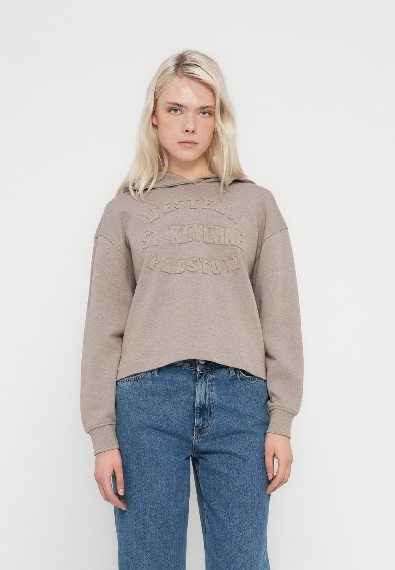 Kratka svetlo rjava trenirka z izbočenim napisom. Ima kapuco z vrvico in sproščene rokave. Kombinirana z modrimi kavbojkami z visokim pasom.