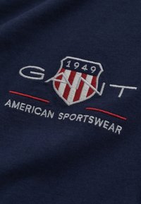 Tissu en coton marine avec un écusson brodé blanc et rouge portant le texte "GANT", "1949" et "AMERICAN SPORTSWEAR" en dessous.