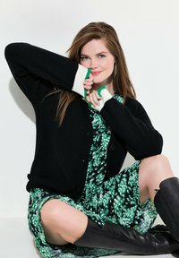 Cardigan noir avec des accents blancs et verts sur une robe florale verte. Modèle assis avec des bottes noires montantes, texture douce et coupe décontractée.