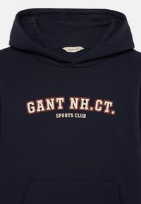 Marinblå hoodie med känguruficka och dragskohuva. Har framträdande vit och röd text: "GANT NH.CT. SPORTS CLUB."