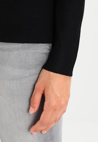 Sort ribbet sweaterærme synligt, parret med lysegrå stof. Håndleddet er eksponeret, hvilket viser glat hud.