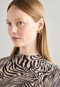 Des boucles d'oreilles circulaires en ton doré présentent un design torsadé. Le sujet porte un haut beige avec un motif zèbre noir. Texture lisse et drapé doux.