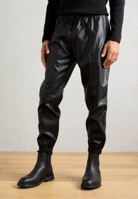 Hombre vestido con pantalones de chándal de imitación cuero negro con puños elásticos y botas de cuero negras hasta el tobillo, de pie sobre un suelo de madera.
