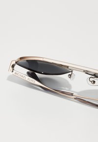 Óculos de sol Michael Kors com armação metálica em ouro rosa, lentes pretas, almofadas nasais transparentes e nome da marca gravado na têmpora.