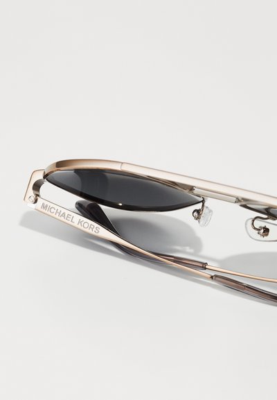 Lunettes de soleil Michael Kors avec monture en métal doré rose, verres noirs, plaquettes de nez transparentes et nom de marque gravé sur la branche.