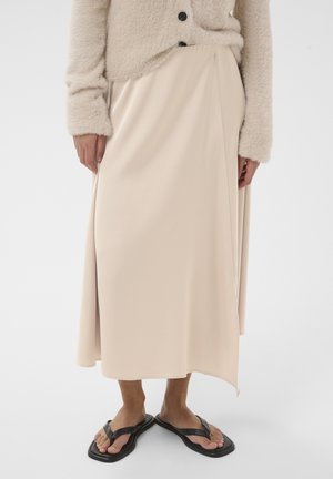 Osoba oblečená v béžovom chlpatom dlhom rukáve, svetlobéžovej midi sukni s bočným rozparkom a čiernych šľapkách so štvorcovými špičkami, stojaca na bielom pozadí.