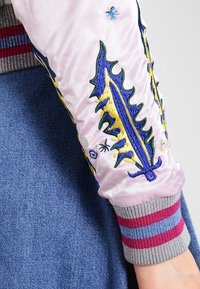 Rękaw jasnoróżowej satynowej kurtki bombowca z kolorowymi haftowanymi wzorami, w tym motywem liścia. Szary ściągacz z różowymi i niebieskimi paskami.