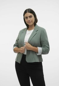 Zelený blejzr s módním střihem, s jednou knoflíkovou úpravou a vyhrnutými pruhovanými manžetami. Nositý přes bílou košili a černé kalhoty.
