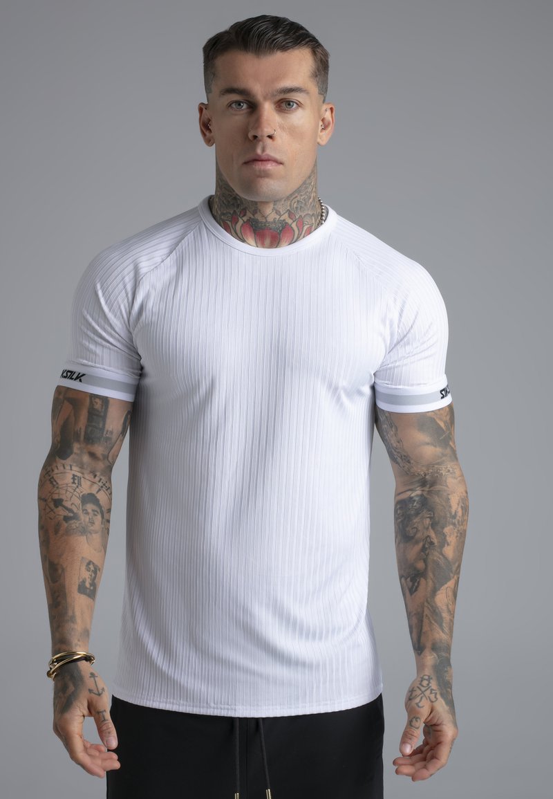 Homme avec des tatouages sur le cou et les bras, portant un t-shirt blanc à manches courtes côtelé et un pantalon noir, debout devant un fond gris.