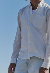 Homme portant une chemise blanche à boutons par-dessus un t-shirt blanc et un jean bleu clair, debout en plein air sous un ciel bleu clair.