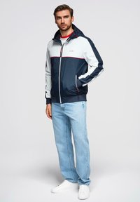 Marineblå og hvid zip-jakke med røde detaljer, udstyret med hætte og sidelommer, parret med lyseblå jeans og hvide sneakers.