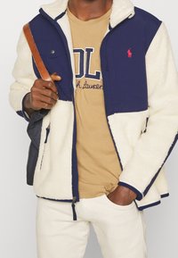 Námořnicky modrá a krémová fleecová bunda se zipovým zapínáním, detaily kapes a červeným logem. Nošena přes hnědé tričko a bílé kalhoty.