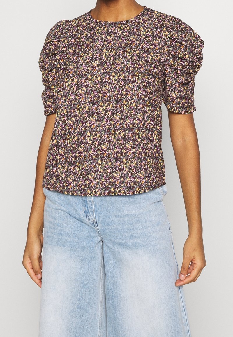 Femme portant un chemisier violet à fleurs à manches courtes avec des épaules bouffantes et un jean large taille haute bleu clair.