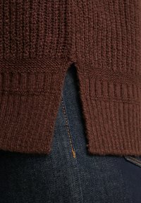 Detail des braunen Strickpullovers mit Schlitzen an den Seiten über dunkelblauen Jeans mit sichtbaren orangefarbenen Nähten.
